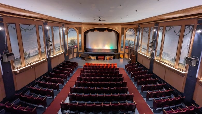 In a large auditorium is a white screen with rows of red chairs. On the walls are panels with murals of trees and the sky. 