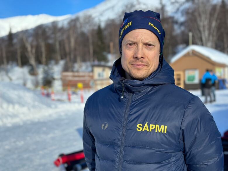 A man in a coat stares straight with a snowy hill behind him. 