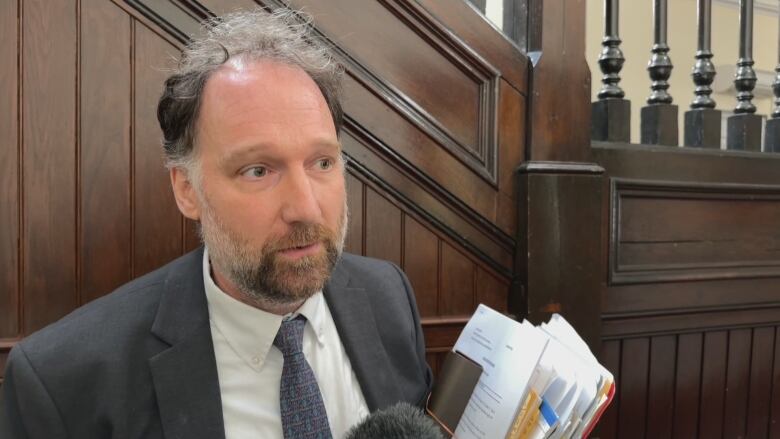 A man in a gray suit and tie speaks to the camera with a brown wooden staircase in the background in the Halifax courthouse. 