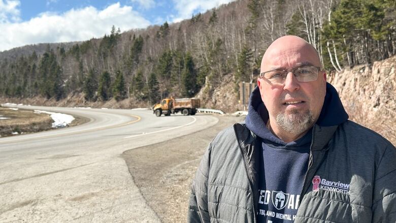 A bald man with glasses and a grey goatee and moustache in a grey puffy jacket stands at the bottom of a curved road with a large yellow highway truck blocking access to a hill.