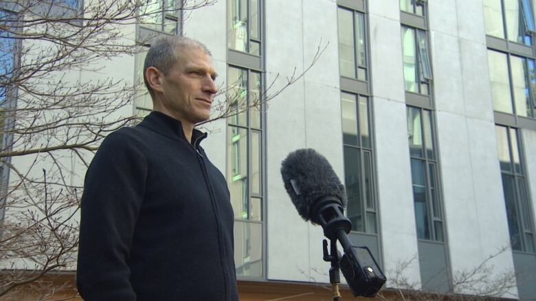 A man with short grey hair and a black sweater stands outside of a building on a university campus.