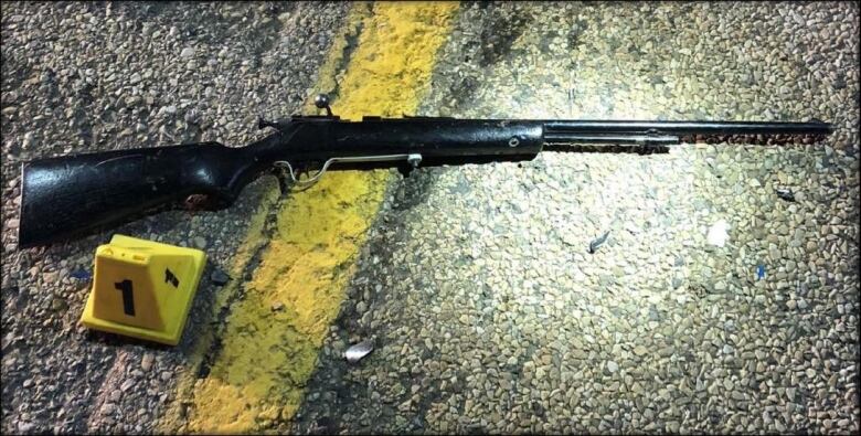 A black rifle sits on a road next to a yellow evidence marker.