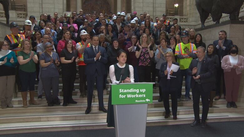 Dozens of people stand on a staircase behind a woman speaking at a podium on the ground.