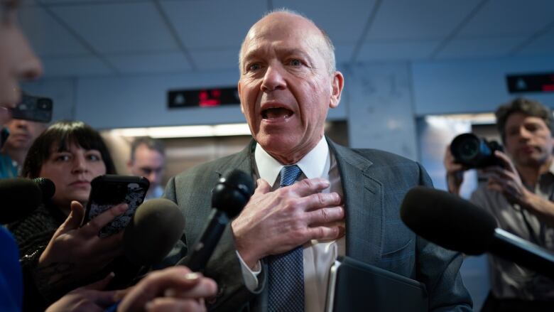 A bald man in a suit, his mouth open mid-sentence and one hand on his chest, surrounded by reporters with microphones and recorders. 