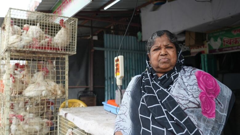 A woman in grey and pink stands for a photo. On the left are chickens in stacked cages.