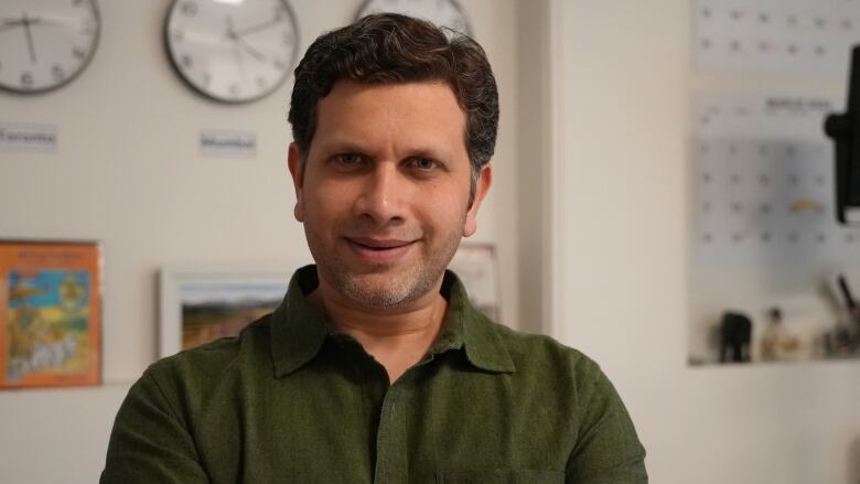 A man in a dark green polo. Behind him are three clocks, some books and two blue calendars.