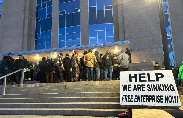 A large group of people standing on a staircase outdoors. A sign in the front reads help we are sinking, free enterprise now. 