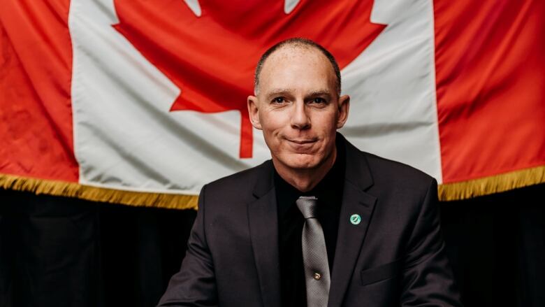 A man in black suit sits at a table with his hand on a bible, with a Canadian flag hung behind him.