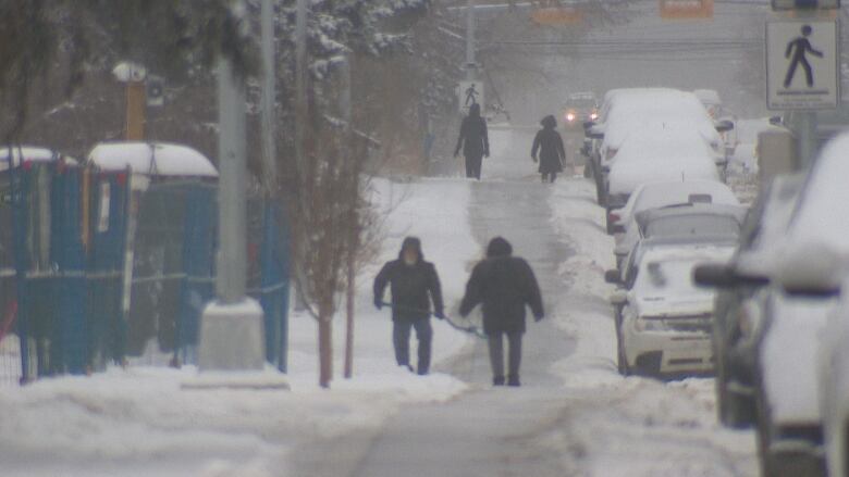 some people walk on a very snowy sidewalk next to cars.