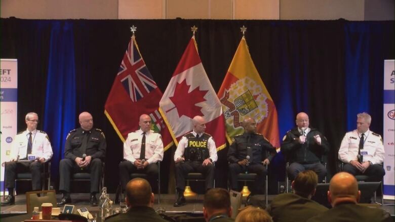 7 police chiefs in uniform sit side by side on a stage. One is speaking into a microphone. There is a crowd in front of them. They are indoors