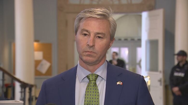 A man stands in a hallway. He is wearing a blue jacket, a white shirt and a green tie.