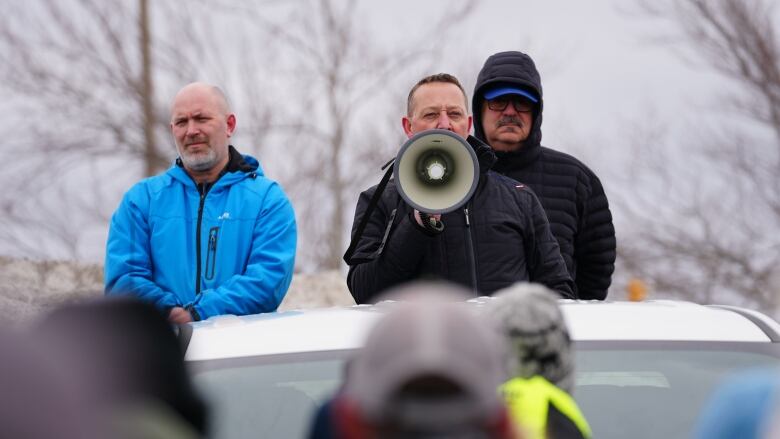 Three men standing in the back of a truck. The guy in the middle is holding a megaphone.