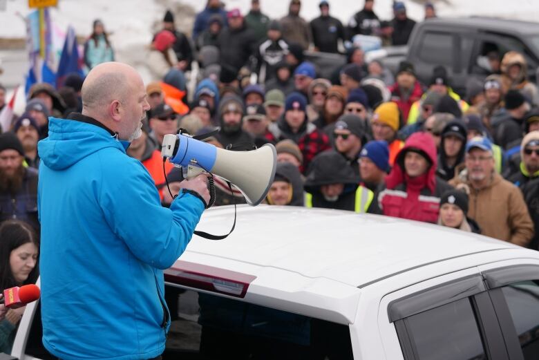 A man in a blue jacket holding a megaphone with a crowd in front of him.