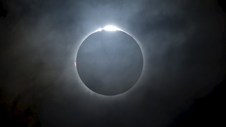 A black disk is seen with thin clouds and thin sunlight peaking out around it.