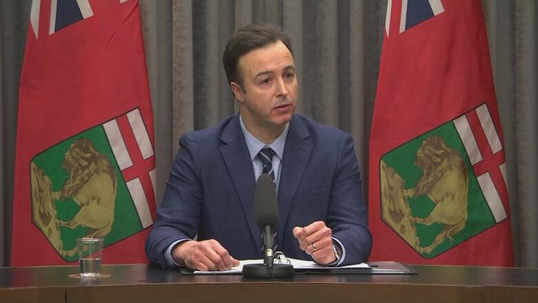 A man in a dark blue suit, light blue shirt and checkered blue tie sits at a table with a microphone in front of him.