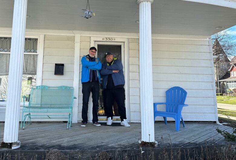 Two men stand on the porch of home. The stand confidently with their arms crossed, and there are to beams supporting an awning in the foreground.