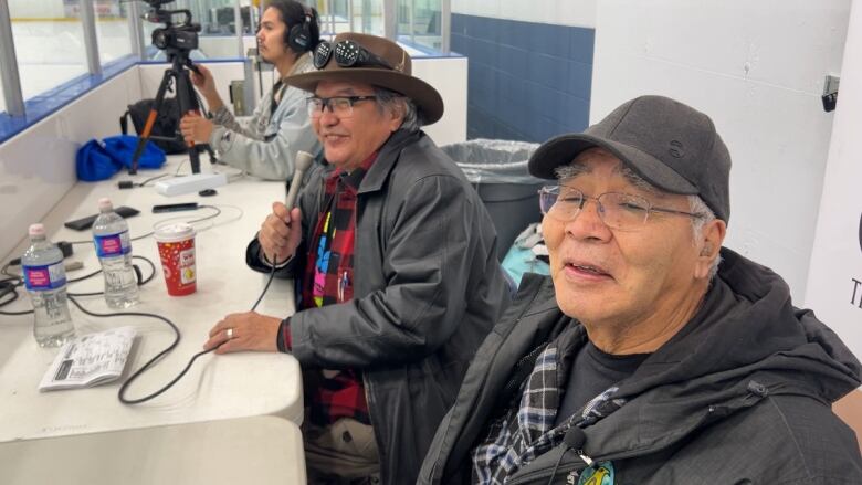 two men sit at a desk beside an ice rink with microphones