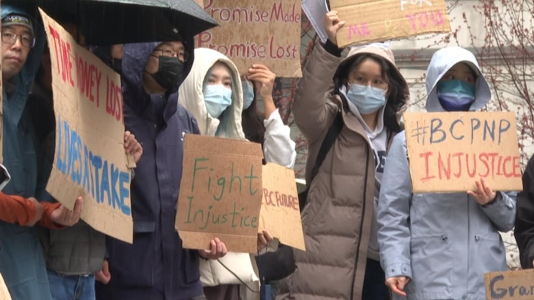 People hold up placards and signs while wearing jackets and facemasks on a rainy day.