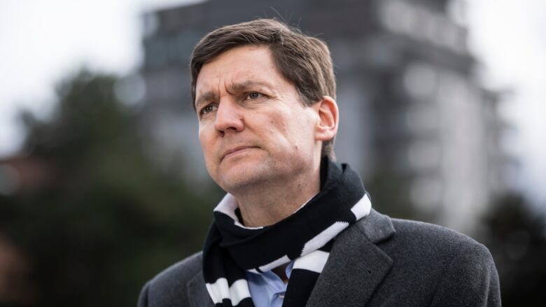 A tall white man wearing a black scarf looks on, with a large building in the background.