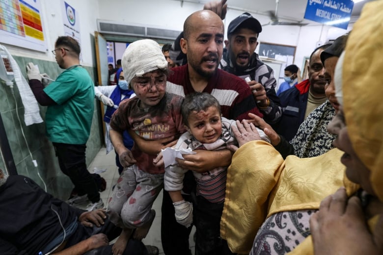 An adult carries injured children inside a crowded hospital.