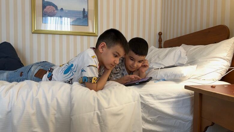 Two young boys watch a tablet while lying on a hotel bed.