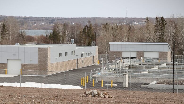 A pair of brown brick buildings with grey trim are shown with a series of pipes and concrete containers on the ground beside them.