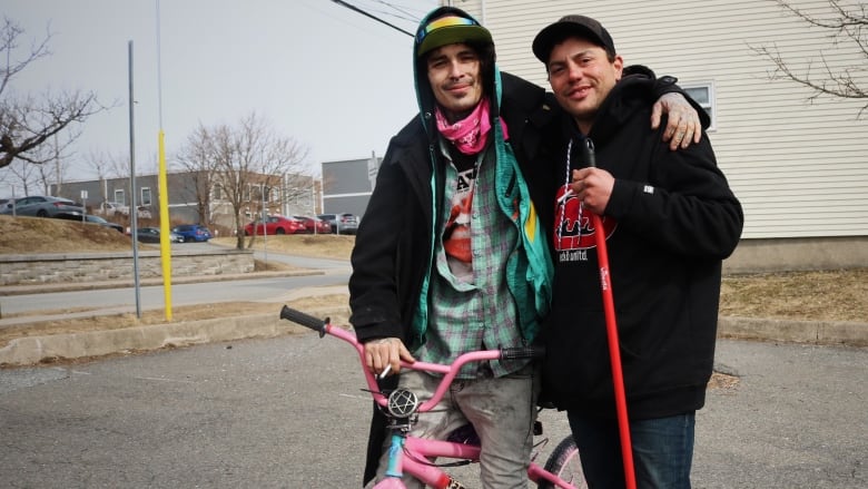 A man on a pink BMX bike poses on the street with a man in a black hoodie holding a red broom. 