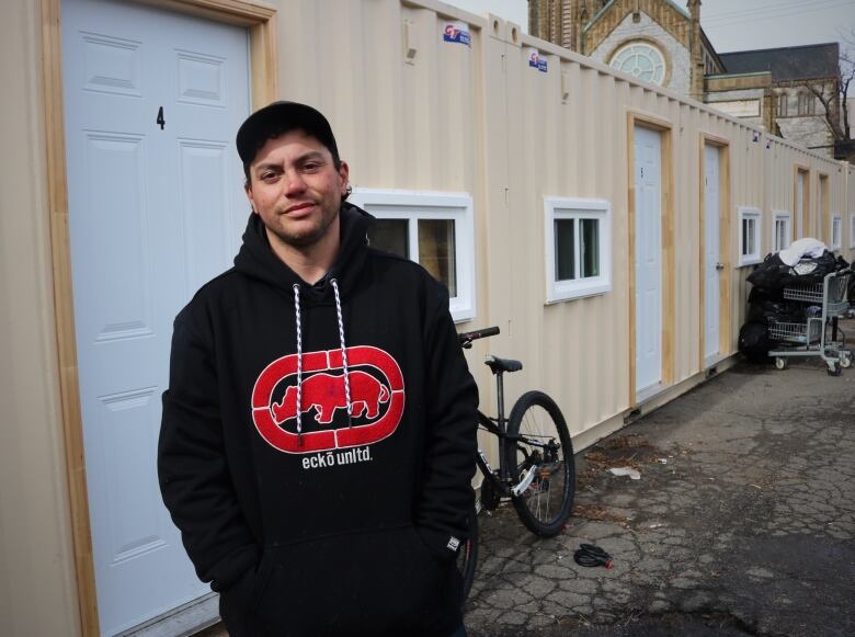 A man in a black Ecko hoodie and a black baseball hat stands in front of shipping containers. 