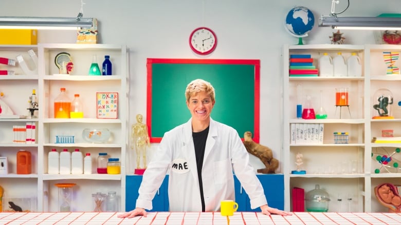 A person with short hair and a lab coat stands at a table in an over-the-top science lab.