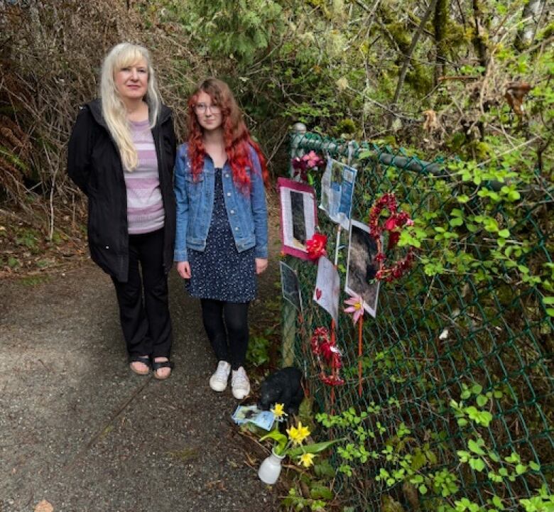 A mother and daughter standing beside a fence inside a park, which serves as a memorial for a dead bear. Photos of the bear, flowers and letters are attached to the fence