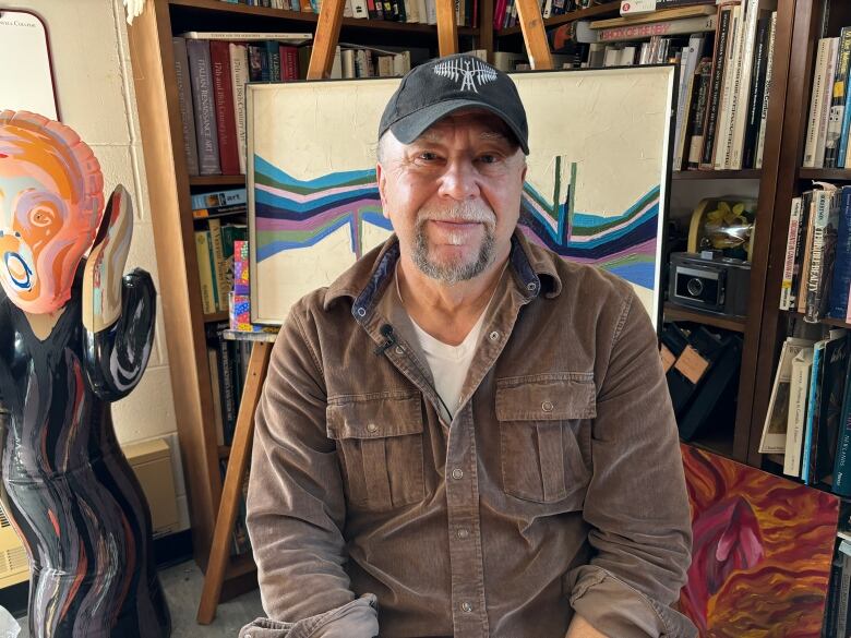 A mean wearing a brown jacket and a cap sits in a artist studio space. An inflatable of the person in The Scream painting stands over his right shoulder.