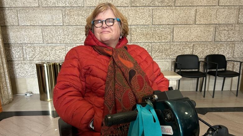 A woman with glasses wearing an orange jacket and orange-patterned scarf is pictured sitting on a mobility scooter in front of a brick wall. 