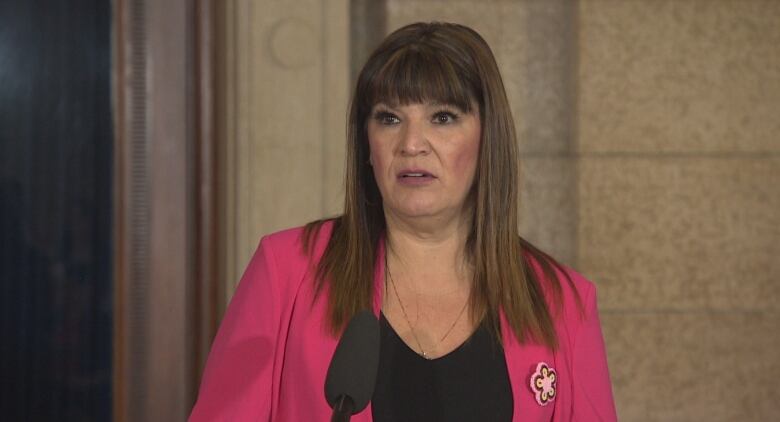 A woman wearing a pink blazer over a black shirt speaks at a microphone.