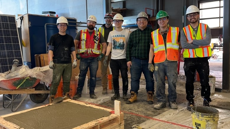 Seven men in hard hats, standing behind a newly-poured concrete slab