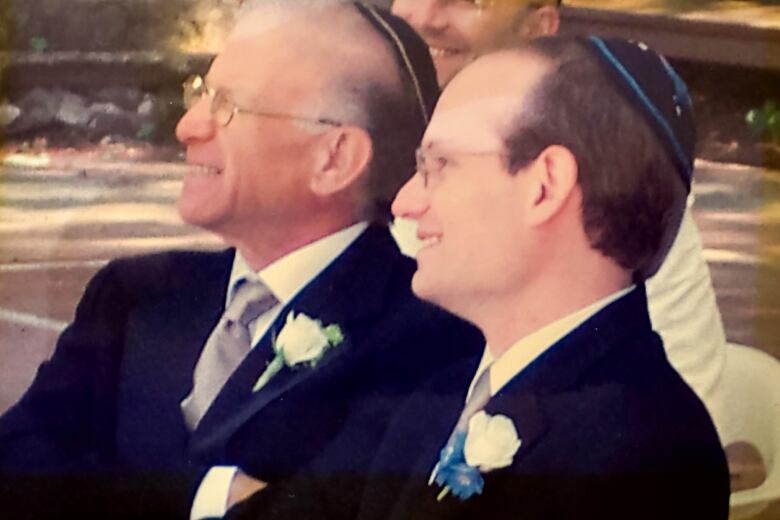 Two men dressed in formal attire sit together and smile at something.