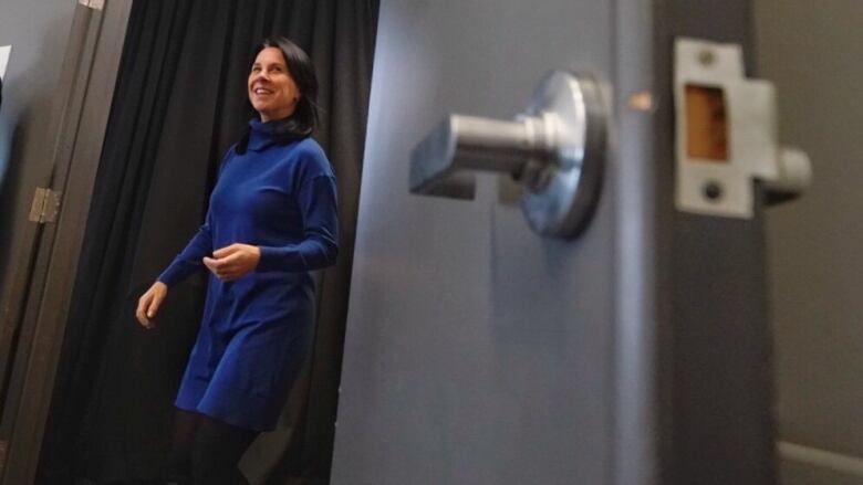 A white woman with dark hair walking through a door, smiling and wearing a blue turtleneck dress.