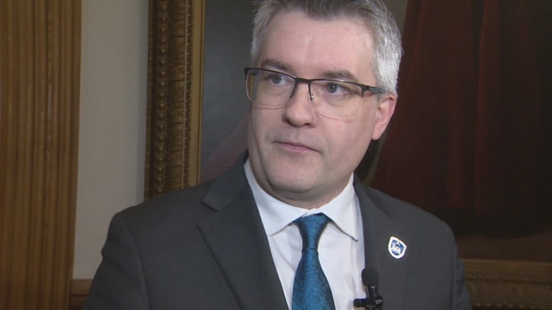 A man in a blue suit and white shirt wearing glasses and a lapel pin.