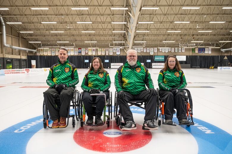 Four people in green uniforms sit in wheelchairs on a curling sheet.