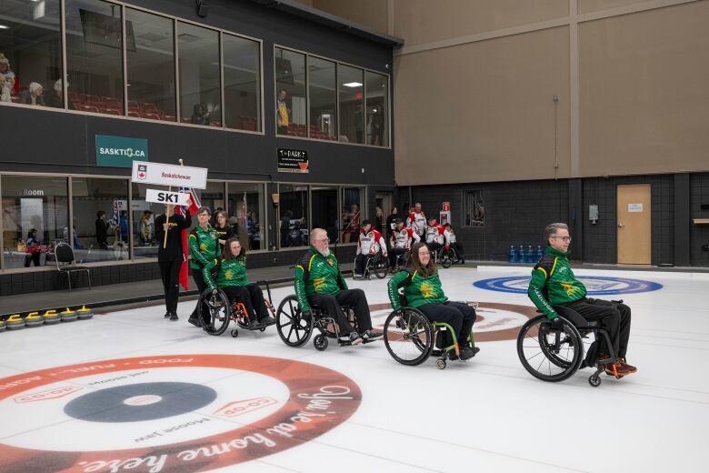 Five people in green uniforms, four in wheelchairs, move across a curling sheet.