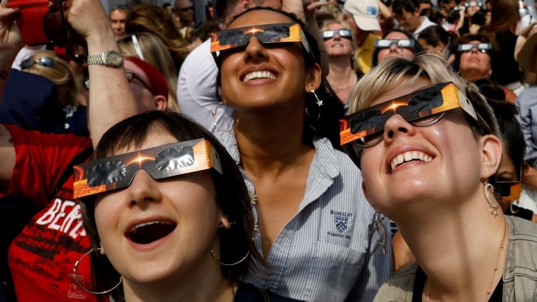 Three young women smiling, wearing eclipse glasses while looking skyward.