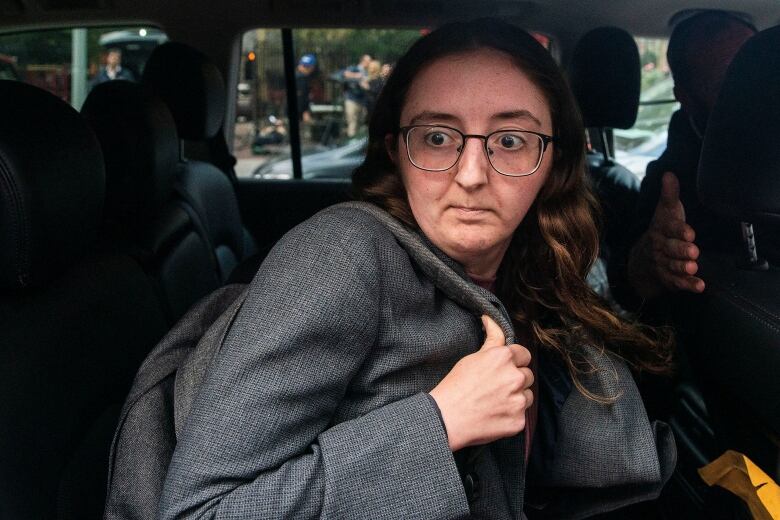 A bespectacled woman is shown in closeup sitting in the back seat of a vehicle.