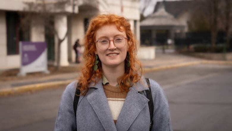 Portrait of a young woman with glasses and curly red hair standing on a street. She is in focus, but the background is out of focus.