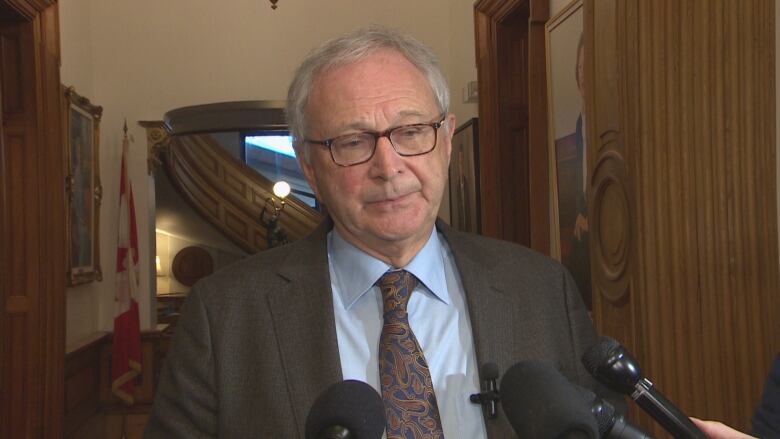 A white-haired man in a suit and tie stands in a hallway talking to reporters.