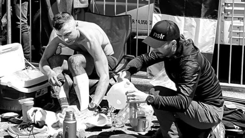 A man is in a chair and is rubbing his foot with a towel. A nother man is crouched nearby filling a water bottle with water. On the floor around them are bottles, shoes, and coolers. 