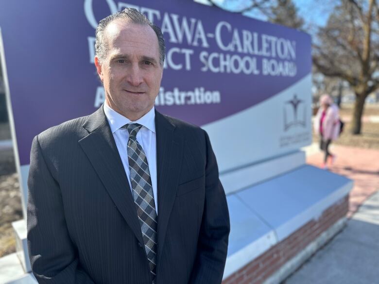 A man wearing a suit stands in front of a sign.