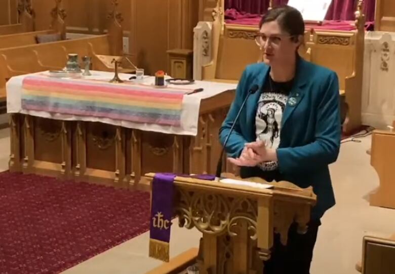 A individual in a blue blazer stands before a pulpit. Behind them is a rainbow flag.