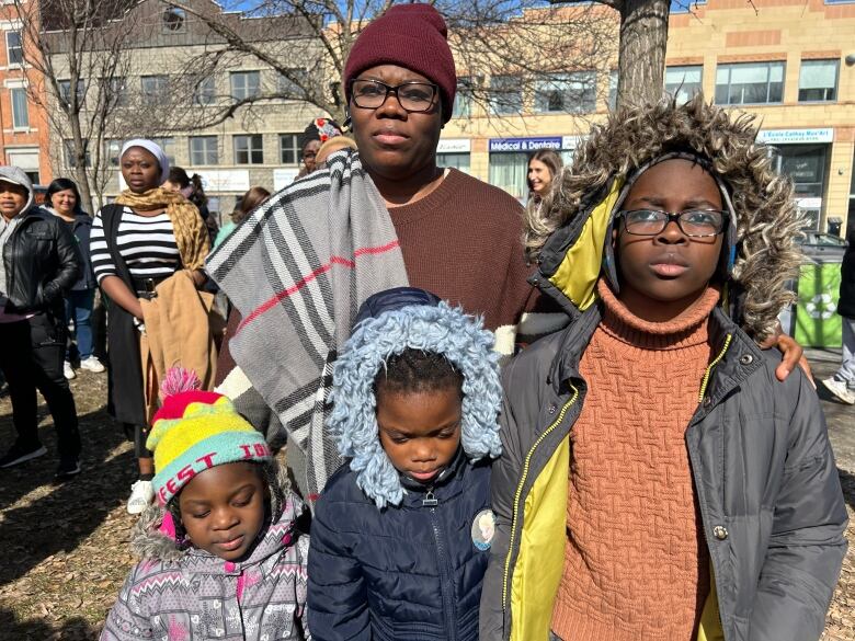 A mother stands behind her three children outside, wearing hats, sweaters and winter jackets. 