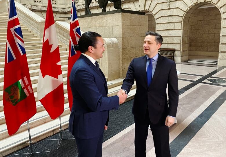 Two men in suits shake hands at the base of a staircase in front of Manitoba and Canadian flags