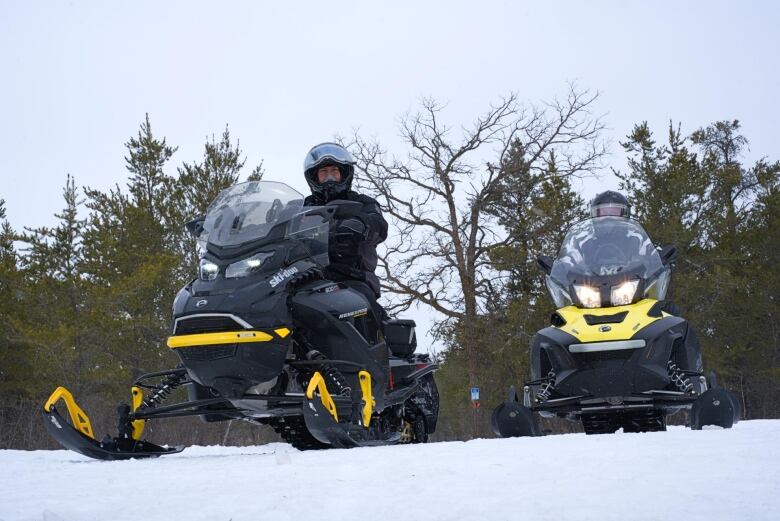 Two men sit on snowmobiles on a snowy trail in the woods.