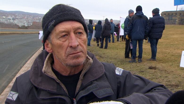 A man wearing a grey jacket and black winter hat stands on a grassy area beside a road.